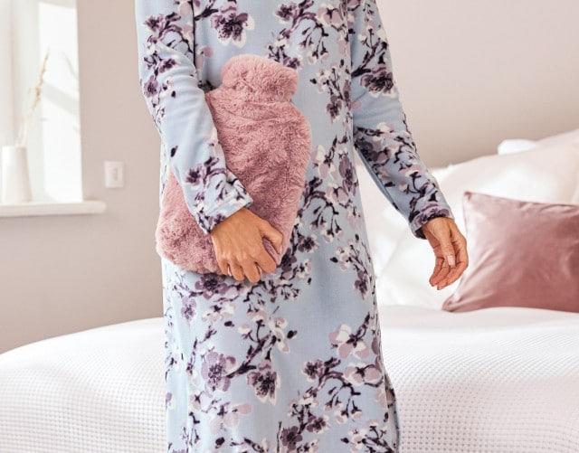 Wärmflasche mit kuscheligem rosa Teddyfleece wird von einer Dame im Nachthemd ins Schlafzimmer getragen.