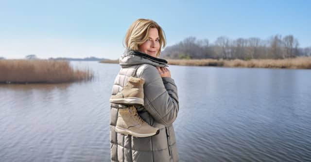 Frau steht am winterleichen See in einer Winterjacke gekleidet und präsentiert neue Winterboots, in dem sie über die Schulter gehangen werden.