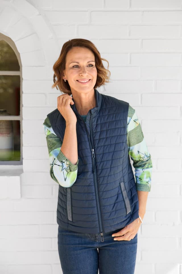 Dame steht vor einer weißen Hauswand und trägt über einem Shirt mit 3/4Arm eine dunkelblaue Steppweste.