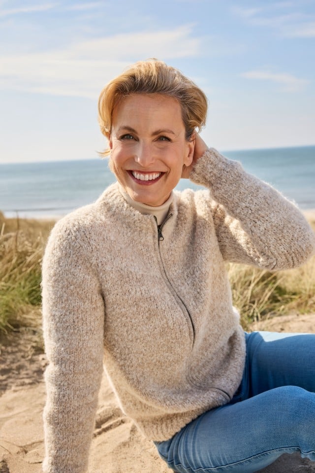 Dame sitzt am Strand und trägt eine kuschelige Strickjacke aus Alpaka.