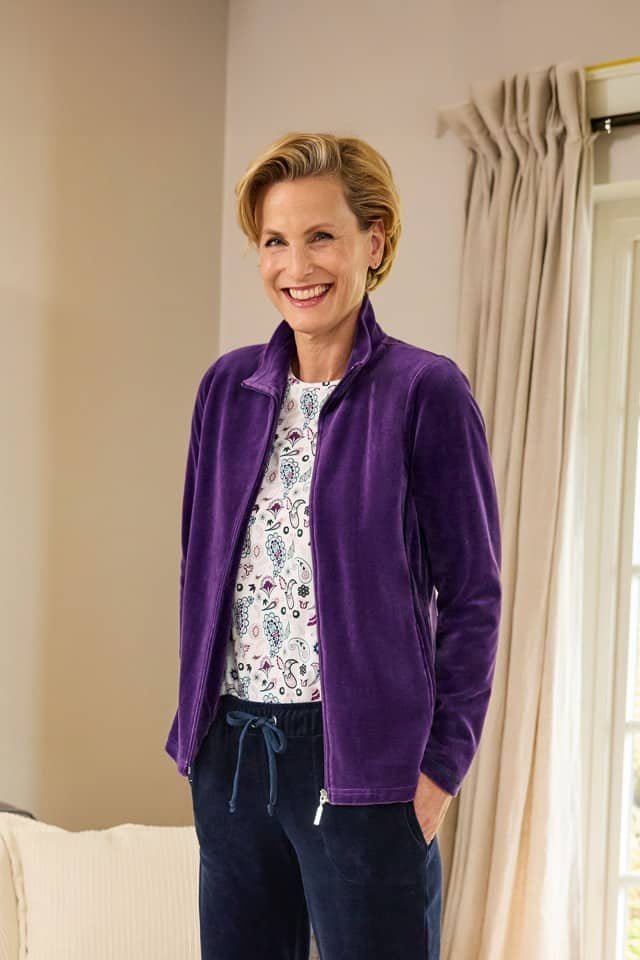 Frau steht im Wohnraum und trägt einen Hausanzug aus violettfarbenem Nickistoff.