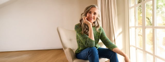 Frau mit grünem Strickpullover sitzt im Sessel an einer großen Fensterfront und die Sonne strahlt in den Wohnraum.