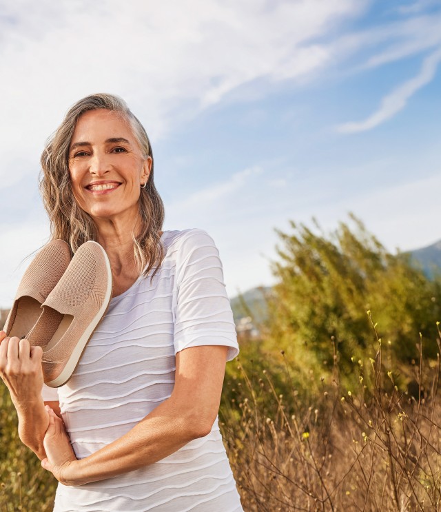 Die grauhaarige Frau genießt das Leben und steht bei sonnigem Wetter mit ihren Hallux-Slipper in der Hand in einer sommerlichen Landschaft.