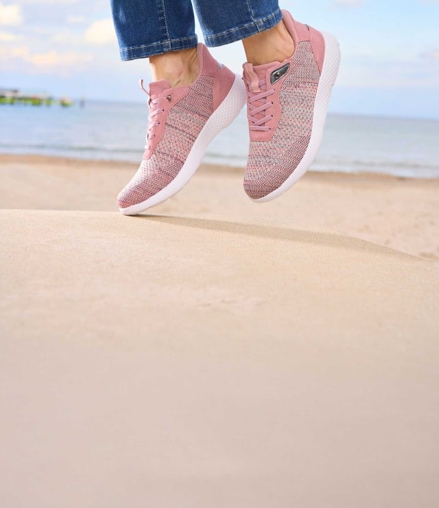 Frau springt am Strand mit den Hallux-Schlupf Schnürer in Rosa.
