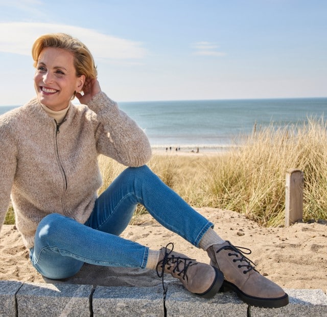 Auf einer Mauer, unmittelbar am Strand, sitzt eine Frau im Sand und trägt die bequemen Hallux-Stiefelette Trittsicher in der Farbe Grau.