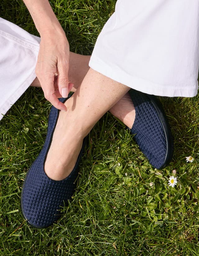 Frau sitzt auf grüner Wiese im Schneidersitz und trägt den Hallux Softslipper in dunkelblau. 
