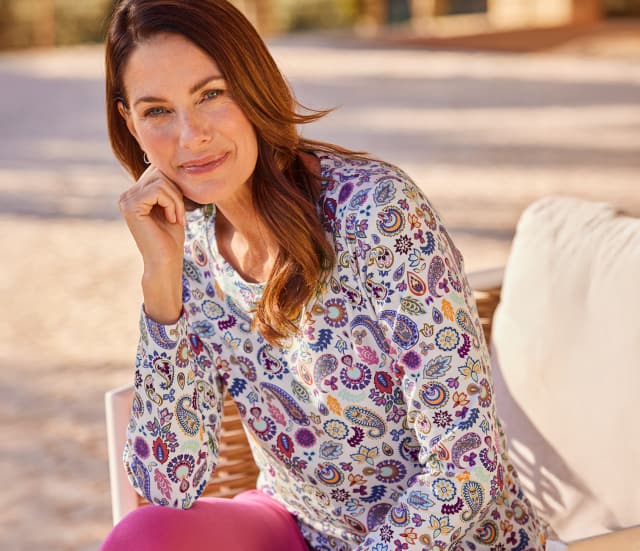 Frau sitzt glücklich in der Sonne auf einer Terrasse und trägt Baumwoll-Shirt Paisley in Beere-Tönen.