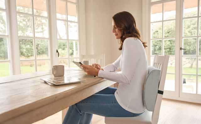 Eine lächelnde Frau sitzt am Tisch und hält ein Tablet in der Hand. Vor ihr, auf dem Tisch, steht eine Tasse.