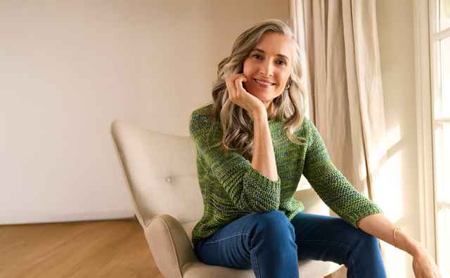 Aeltere Frau sitzt lächelnd in einem Sessel am Fenster, trägt grünen Pullover und blaue Hose. Der Raum ist hell mit Holzboden und Vorhängen.