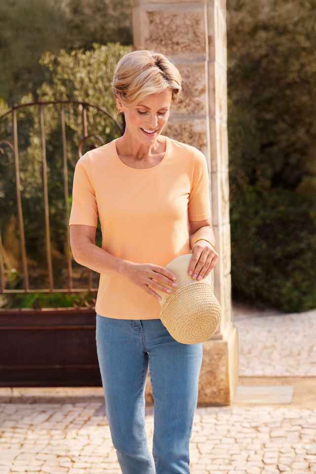 Frisches pfirsichfarbenes Shirt trägt eine Frau zur hellblauen Jeans und hält einen Sommerhut in den Händen