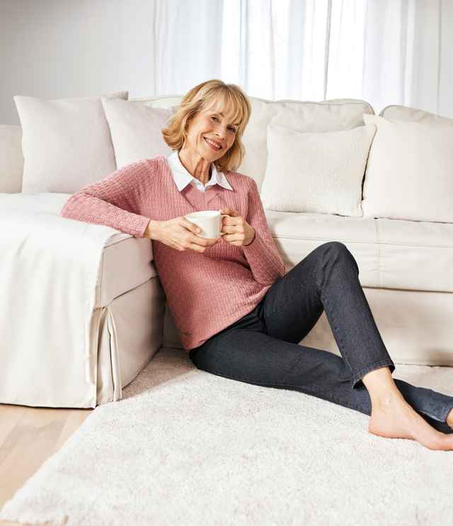 Lächelnde Frau sitzt angelehnt an einer Couch und trägt eine dunkle Jeans.
