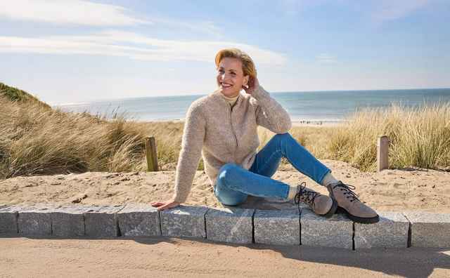 Frau sitzt lächelnd auf einer Mauer am Strand, trägt Pullover und Jeans. Im Hintergrund Sanddünen, Meer und blauer Himmel.