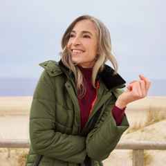 Frau in grüner Jacke lächelt und steht vor einem Strandhintergrund. Sie hat graues Haar und trägt einen roten Pullover unter der Jacke.