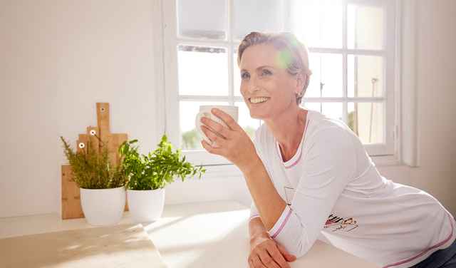 Frau in weißem Shirt trinkt Kaffee in heller Küche, lächelt und lehnt sich auf Tisch. Pflanzen und Fenster im Hintergrund.