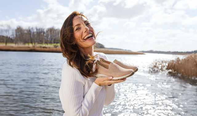 Eine lachende Frau in weißem Pullover steht am Wasser und hält beige Schuhe in der Hand. Im Hintergrund blauer Himmel und Bäume.