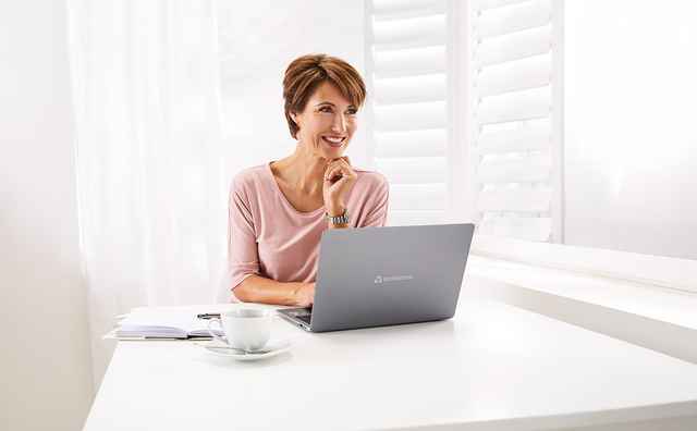 Frau in rosa Bluse sitzt lächelnd an einem weißen Tisch mit Laptop, Notizbuch und Kaffeetasse vor hellen Fensterjalousien.