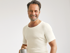 Lächelnder Mann mit grauen Haaren in einem weißen T-Shirt hält eine graue Tasse vor einem schlichten Hintergrund.