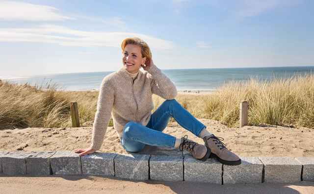 Eine Person sitzt lächelnd auf einer Steinmauer am Strand, trägt einen Pullover und Jeans, mit Gras im Hintergrund und Meer im Hintergrund.