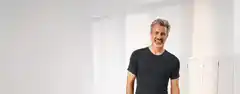 Ein Mann mit grauem Haar und Bart trägt ein dunkles T-Shirt und steht lächelnd in einem hellen Raum mit Vorhängen und Fensterläden.