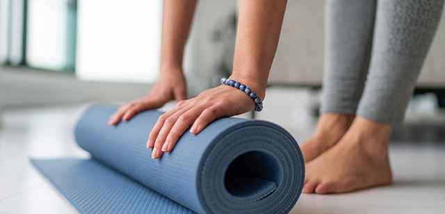 Nahaufnahme von Händen, die eine blaue Yogamatte auf einem weißen Boden ausrollen. Eine Person steht barfuß daneben.