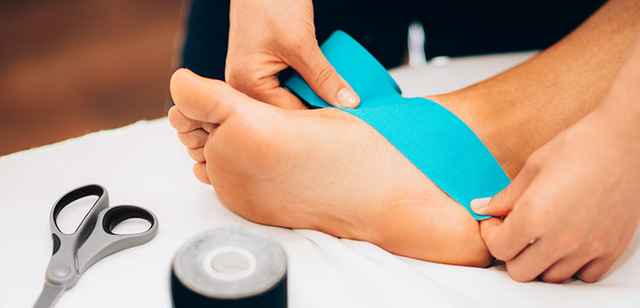Person legt einen blauen Kinesiotape-Streifen auf den Fuß eines anderen. Eine Schere und eine Tape-Rolle liegen daneben.
