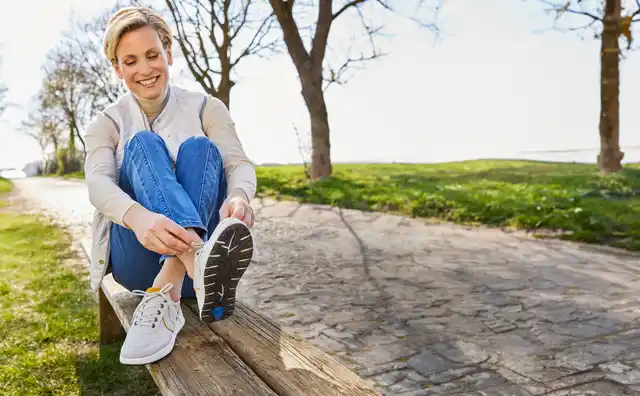 Frau sitzt auf einer Bank im Park und bindet ihre Schnürsenkel. Sie lächelt und strahlt Zufriedenheit aus. Bäume im Hintergrund.