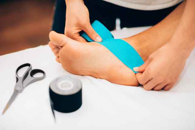 Jemand klebt ein blaues Kinesiotape auf einen Fuß. Daneben liegen eine Schere und eine schwarze Tape-Rolle auf einem Tisch.