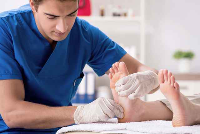 Ein Mann in blauer Kleidung führt eine Fußpflege an einer liegenden Person durch, während er weiße Handschuhe trägt.