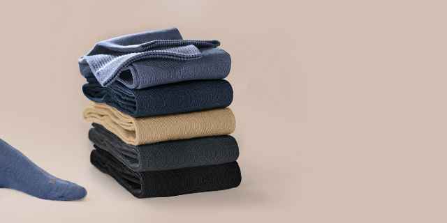 Dunkelblaue Socken an Füßen neben einem Stapel gefalteter Kleidung in verschiedenen Farben auf beigem Hintergrund.
