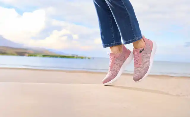 Person in rosa Sneakers springt am Strand; Jeanshose und Meer im Hintergrund.