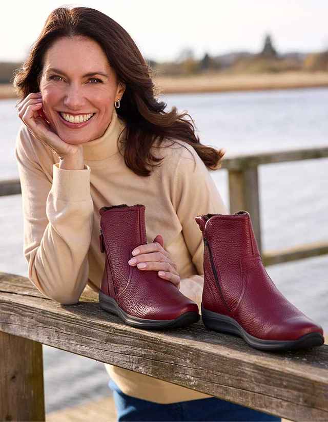 Frau lehnt sich auf ein Geländer und präsentiert lächelnd rote Stiefeletten. Hintergrund zeigt ein Gewässer und unscharfe Landschaft.