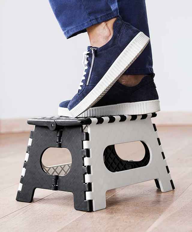 Person steht auf einem kleinen, schwarz-weißen Klapptritt aus Kunststoff, trägt blaue Jeans und blaue Sneakers.