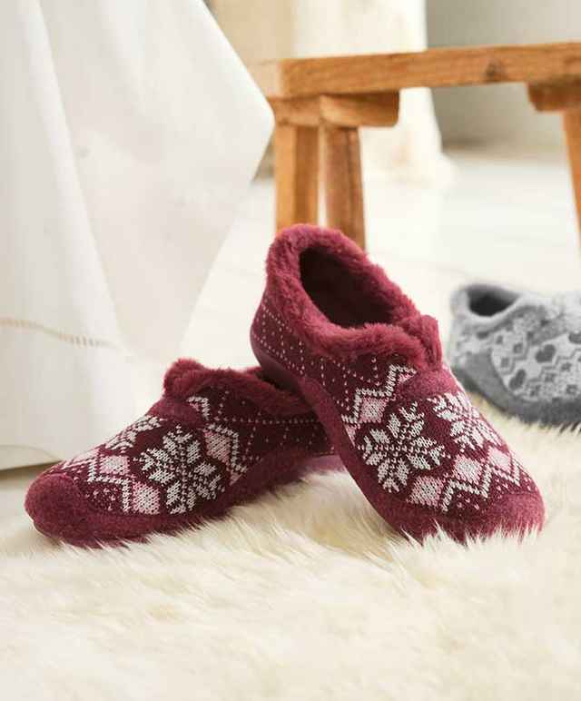 Rote Hausschuhe mit Wintermuster auf flauschigem Teppich neben einem Holzstuhl. Im Hintergrund sind graue Hausschuhe zu sehen.