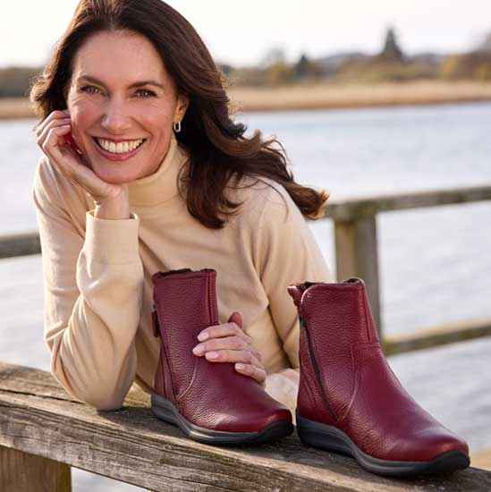 Frau in beige Rollkragenpullover lehnt sich lächelnd auf ein Geländer und hält rote Stiefeletten, Hintergrund ist eine Flusslandschaft.