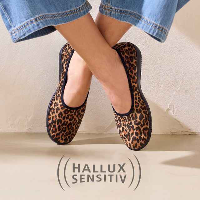 Beine gekreuzt, getragen werden Leopardenmuster-Schuhe mit schwarzem Rand. Text "Hallux Sensitiv" am Boden. Wand im Hintergrund.
