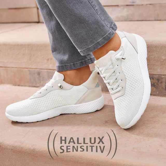 Weiße Hallux-sensitiv Sneaker an Füßen einer Person in grauen Jeans auf einer Steintreppe.