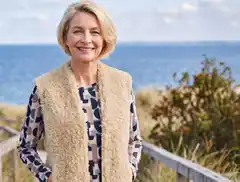 Frau mit blonder Bob-Frisur trägt eine flauschige Weste und lächelt am Strand mit Meer im Hintergrund.