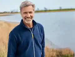 Mann in blauer Jacke lächelt am Seeufer mit Gras im Hintergrund.