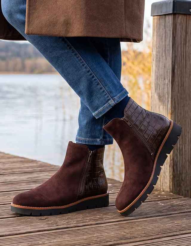 Person trägt braune Stiefel und blaue Jeans, steht auf einem Holzsteg nahe eines Sees im Freien.