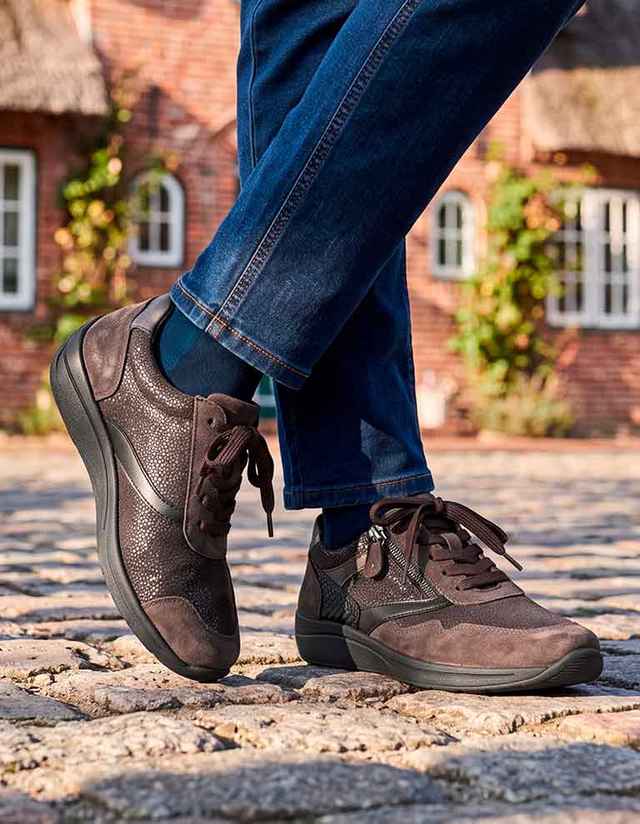 Person trägt braune Sneaker und blaue Jeans, steht auf Kopfsteinpflaster mit Backsteinhäusern im Hintergrund.