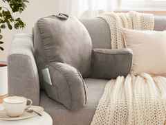 Graues Kissen auf einem Sofa, umgeben von einer cremefarbenen Decke und einem Kissen. Eine Kaffeetasse steht auf dem Tisch daneben.