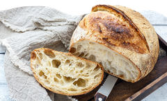Ein Laib knuspriges Brot, teils geschnitten, liegt auf einem Holzbrett mit Messer, daneben ein beigefarbenes Tuch.