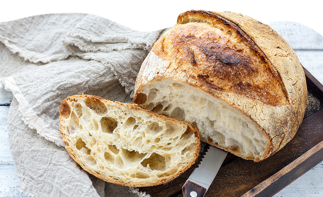 Ein Laib knuspriges Brot, teils geschnitten, liegt auf einem Holzbrett mit Messer, daneben ein beigefarbenes Tuch.
