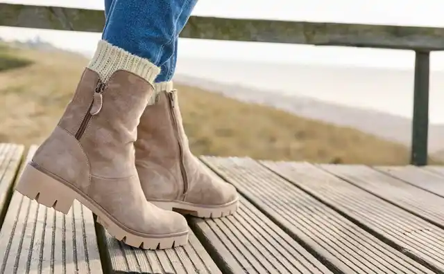 Beine einer Person in beigen Stiefeln und blauen Jeans auf einem hölzernen Steg mit unscharfem Hintergrund einer Küstenlandschaft.