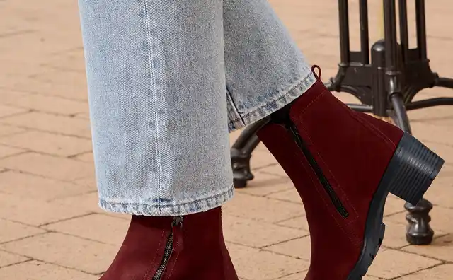 Braune Stiefeletten aus Wildleder mit Reißverschluss und Absatz, kombiniert mit hellblauen Jeans und einem grauen Mantel auf gepflastertem Boden.