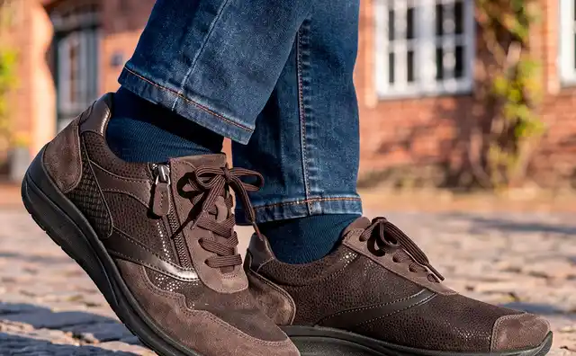 Person trägt braune Sneaker mit Reißverschluss und Jeans auf einer Kopfsteinpflasterstraße vor einem Backsteinhaus.