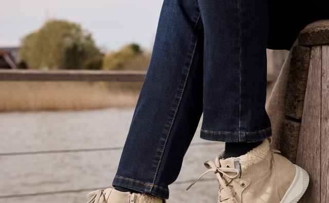 Ein Person trägt beige Sneaker und dunkle Jeans, sitzt entspannt auf einem Holzsteg am Wasser.