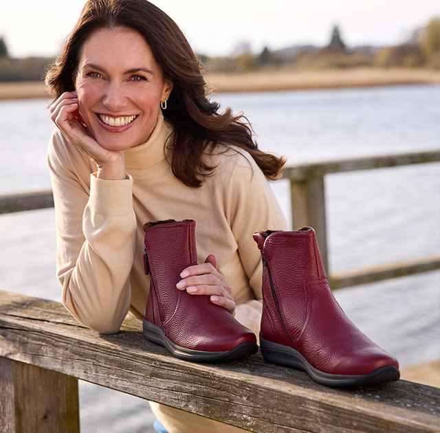 Frau lehnt lächelnd an einem Holzzaun, auf dem ein Paar rotbraune Stiefeletten steht. Im Hintergrund ist ein See zu sehen.